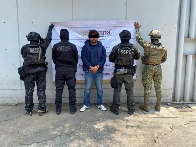 Persona detenida presentada por autoridades federales ante una manta de la Marina